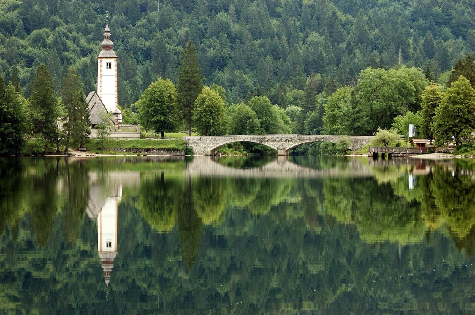 The medieval stone bridge and Gothic tower of Saint John the Baptist church reflected in the mirror-smooth waters of Lake Bohinj