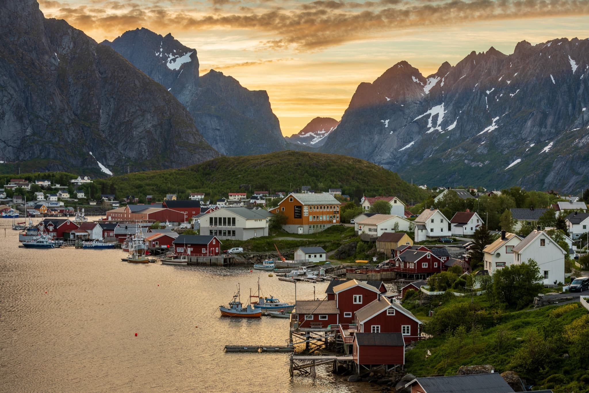 Traditional red wooden rorbuer cabins nestled beneath towering granite peaks along the fjord in Reine, Lofoten